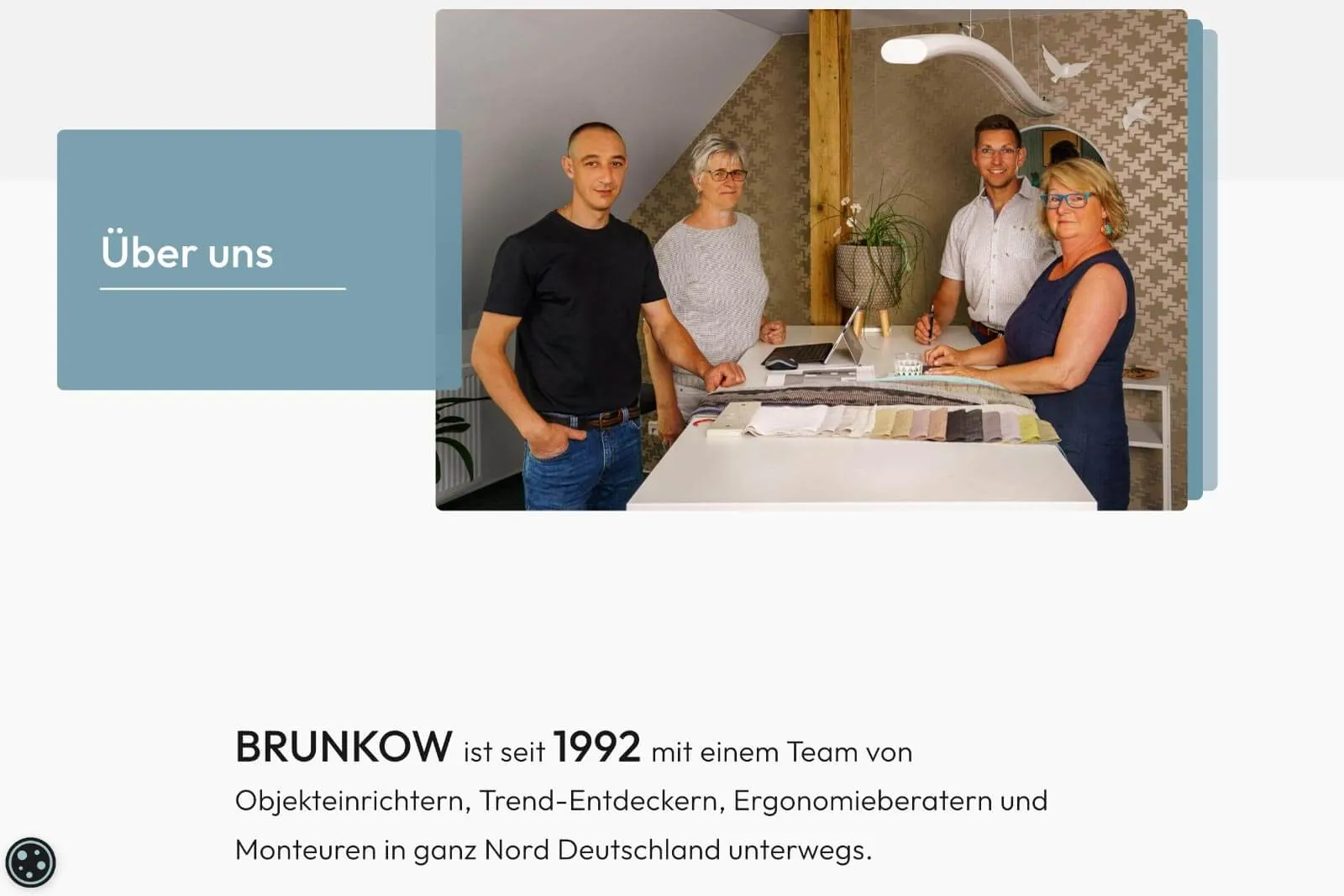 Vier Personen an einem Tisch mit Stoffmustern vor einer Tapetenwand mit Spiegel und Lampe.